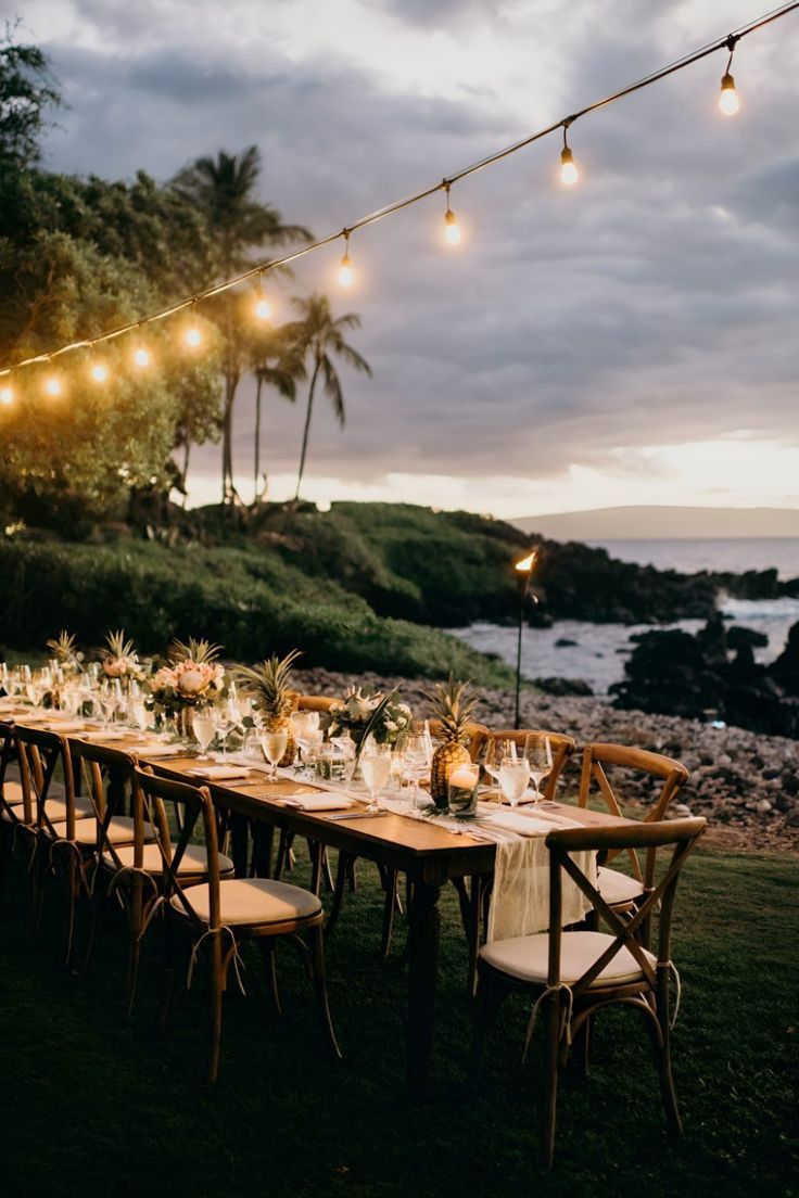 a long table is set up for an outdoor dinner by the ocean with string lights strung over it