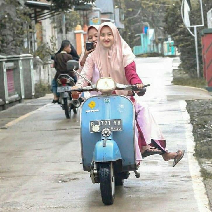 two women riding on the back of a blue scooter down a city street