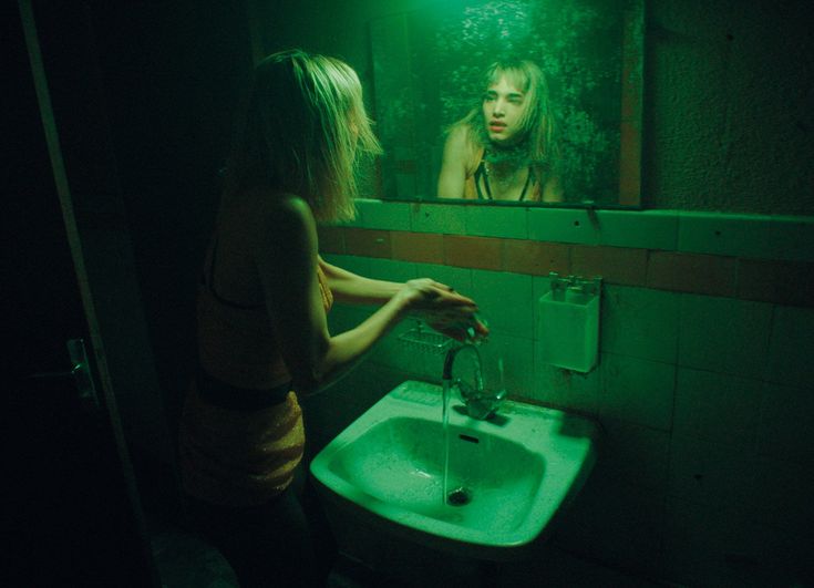 a woman standing in front of a bathroom sink under a mirror with green light shining on her