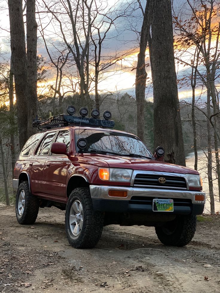 a red truck parked on top of a dirt road in the woods with trees behind it