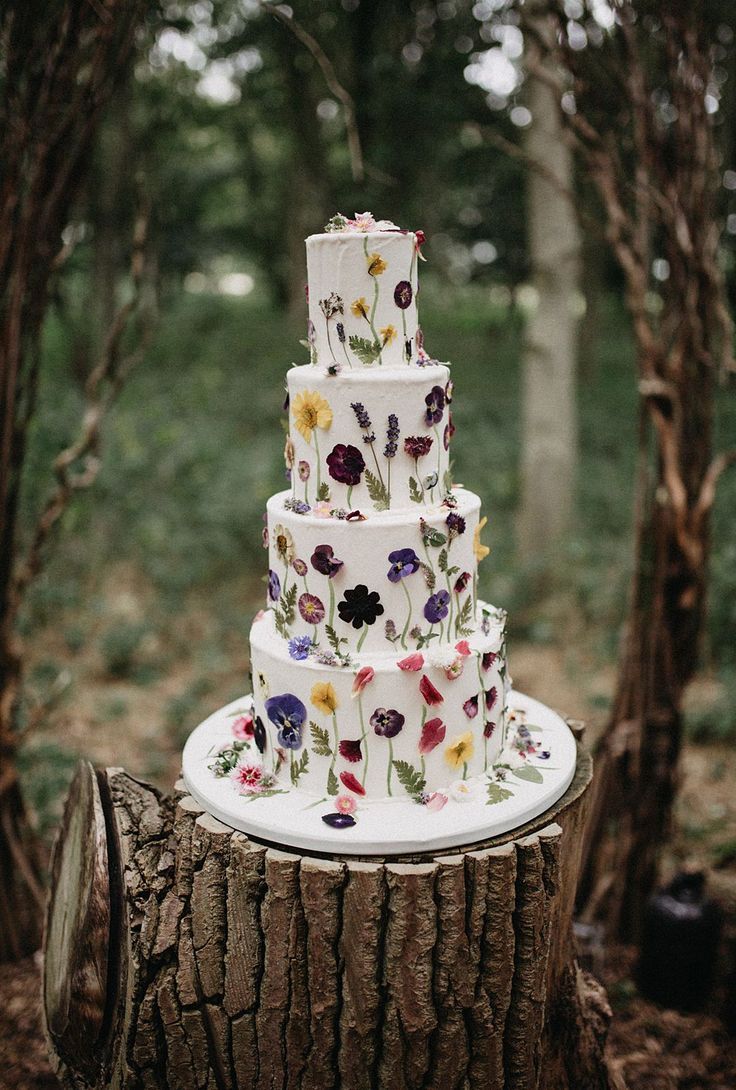 a multi layer cake sitting on top of a tree stump in the woods with flowers all over it