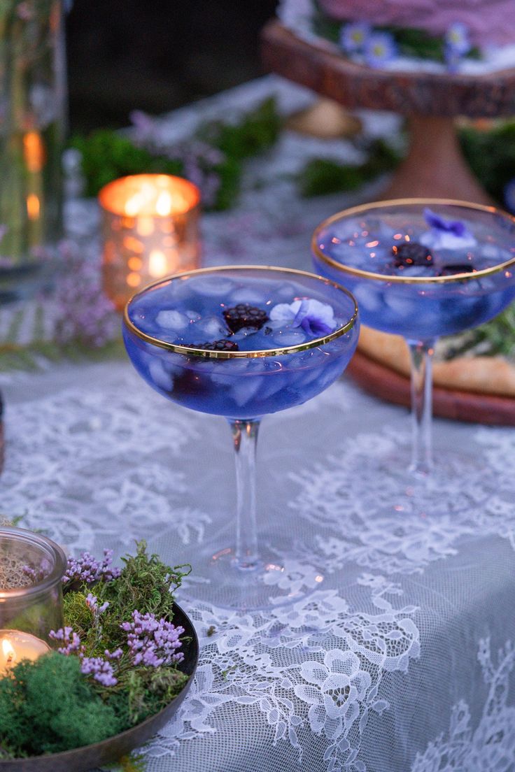 two martini glasses sitting on top of a table covered in purple flowers and greenery