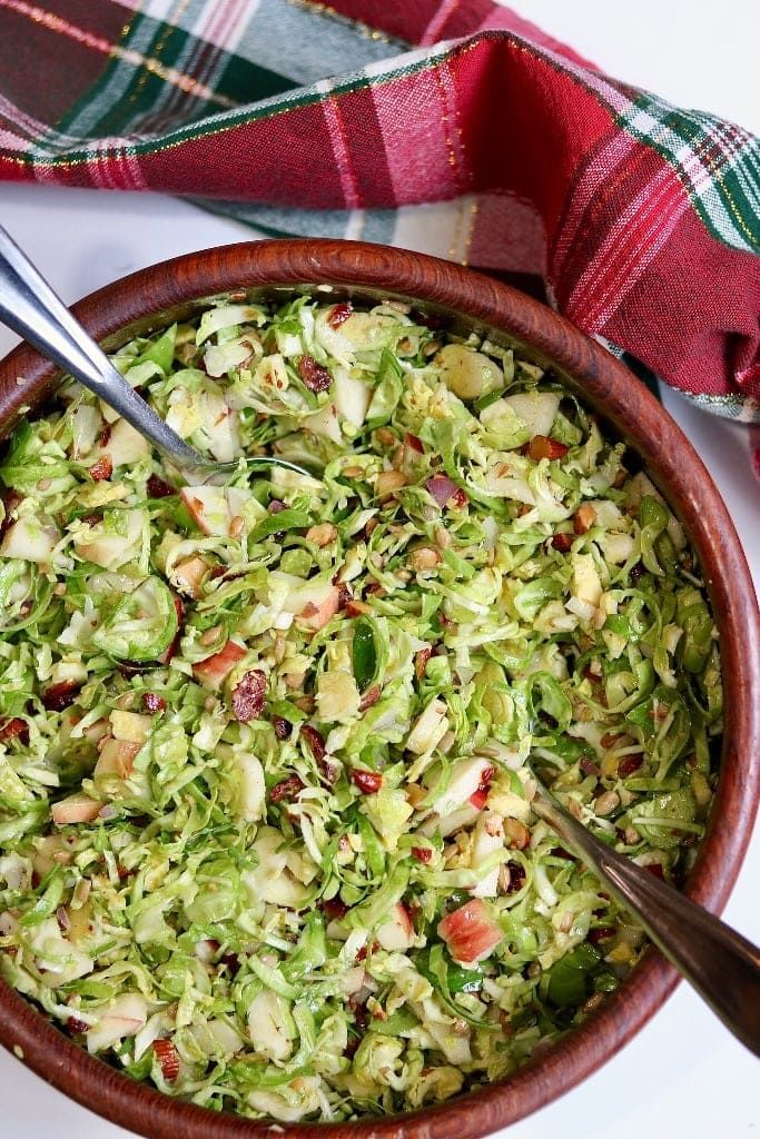 a wooden bowl filled with shaved brussels sprouts and cranberry bits next to a plaid napkin