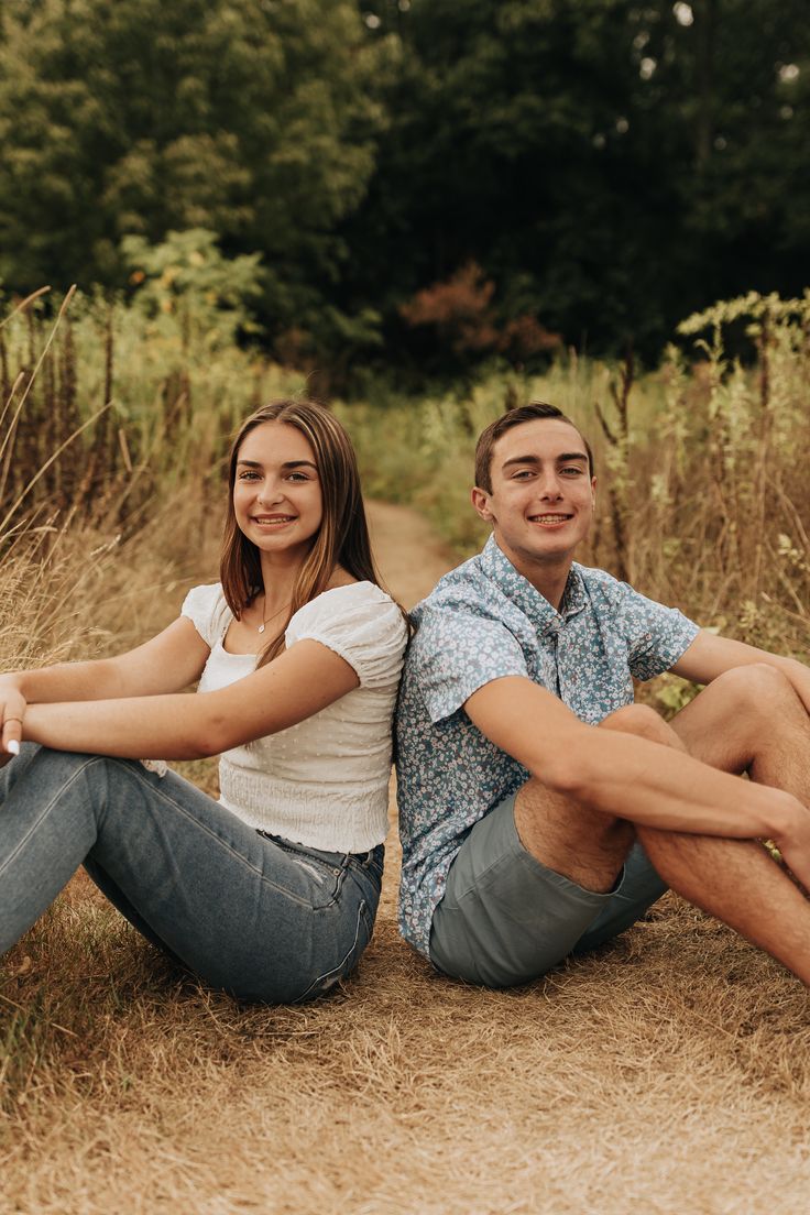 a man and woman sitting on the ground in front of tall grass with trees behind them