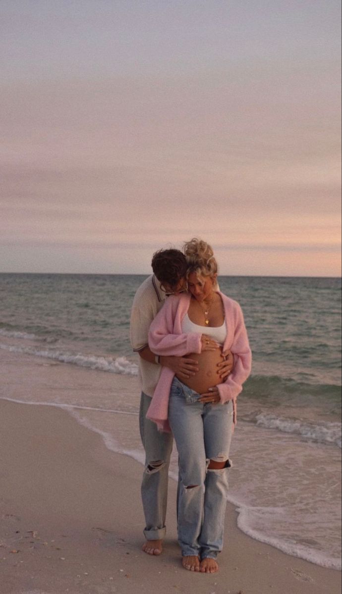 a pregnant woman hugging her husband on the beach at sunset, with waves coming in