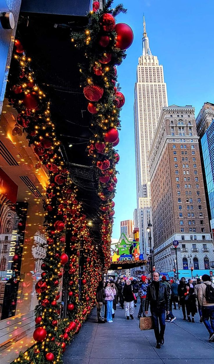 people walking down a street with christmas decorations on the wall and buildings in the background