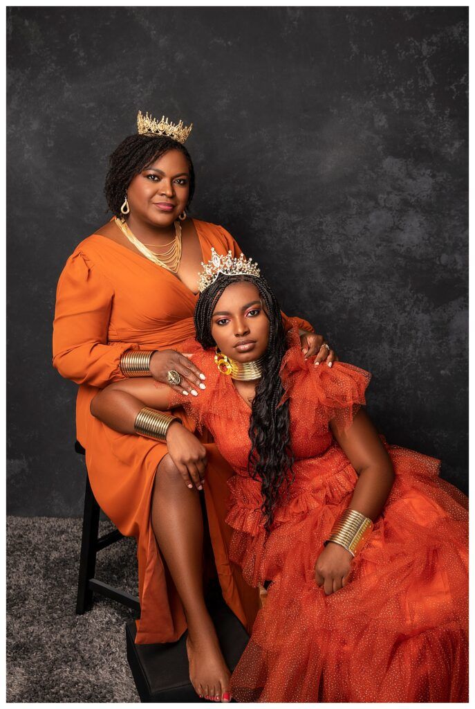 two women in orange dresses and tiaras pose for a photo on a black background