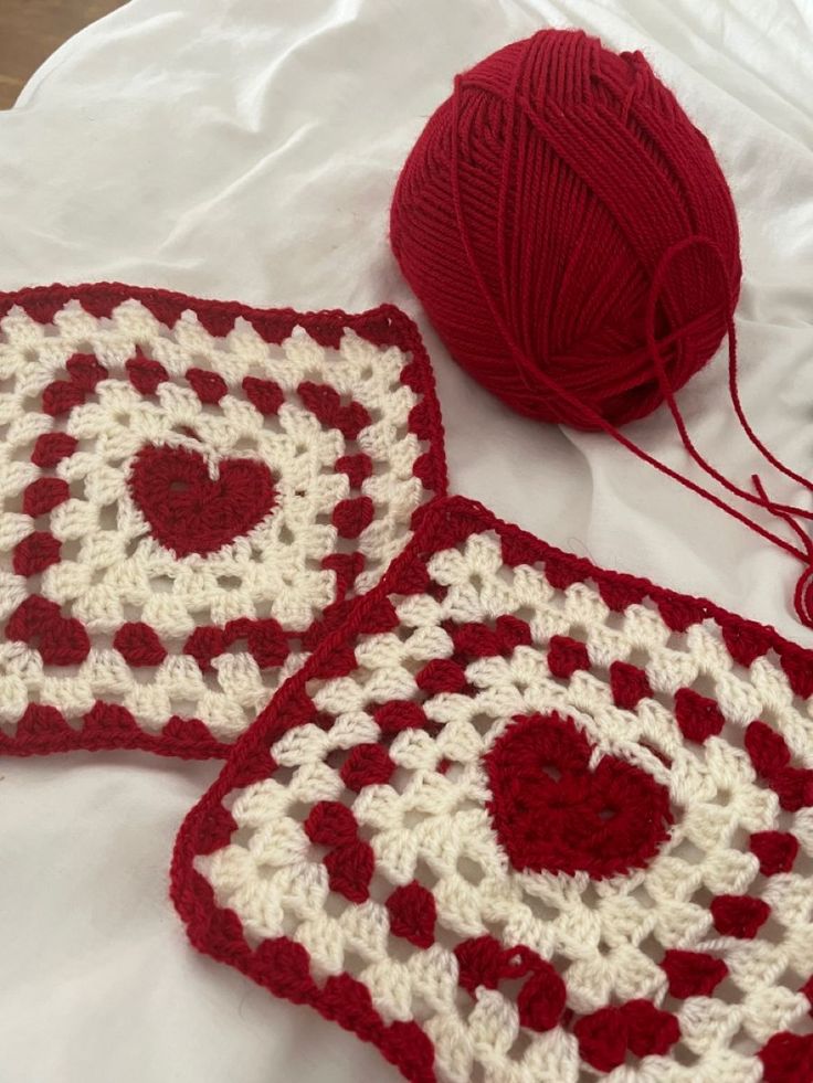 two red and white crocheted squares sitting on top of a bed next to a ball of yarn