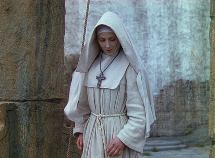 a woman in a nun outfit holding a rope and looking at the camera while standing next to a stone wall