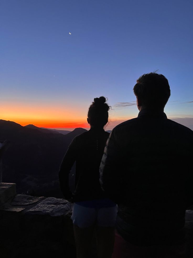 two people standing on top of a mountain watching the sun go down in the distance