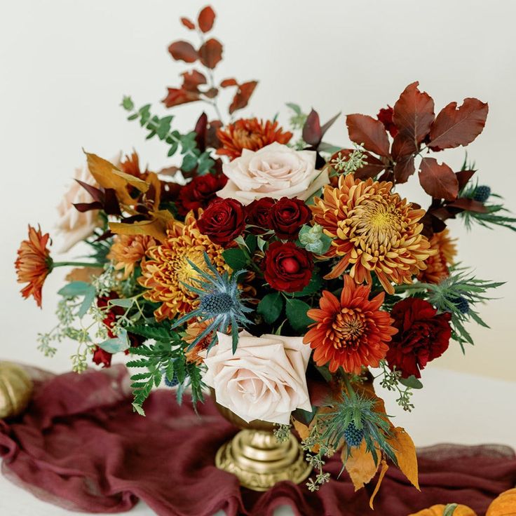 a vase filled with lots of flowers on top of a red table cloth next to pumpkins