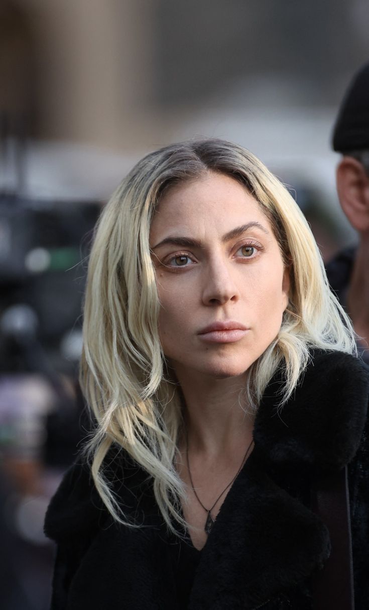 a woman with blonde hair wearing a black coat and looking off to the side while standing in front of a camera