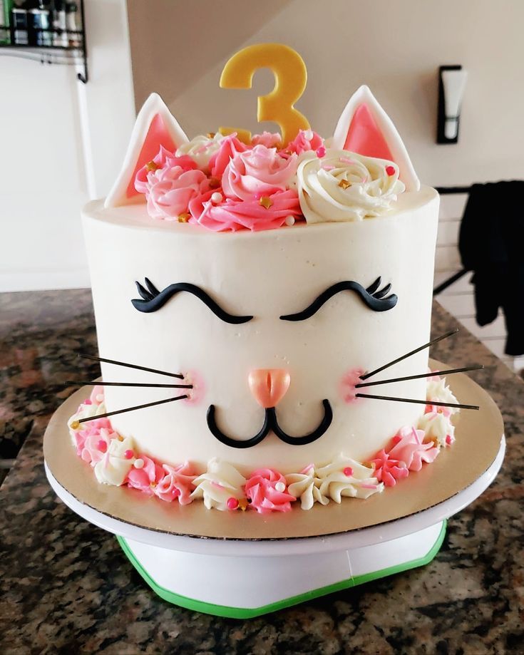 a white cake with pink flowers and a cat face on top is sitting on a counter