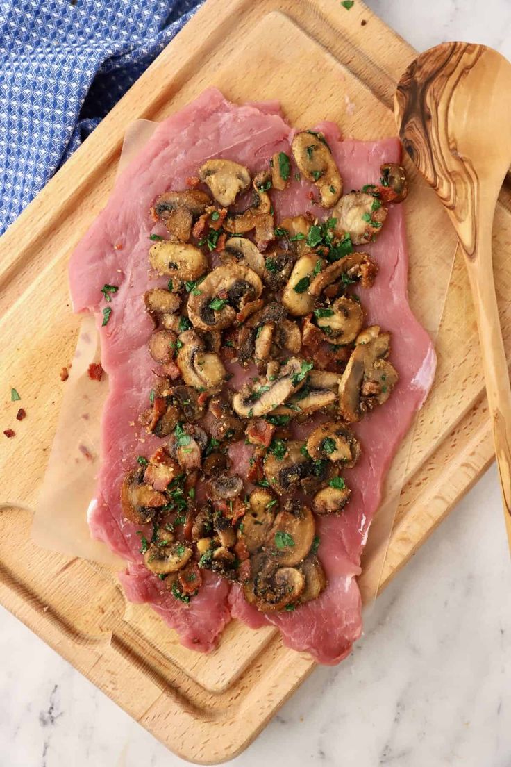 a piece of meat with mushrooms on it sitting on a cutting board next to a wooden spoon