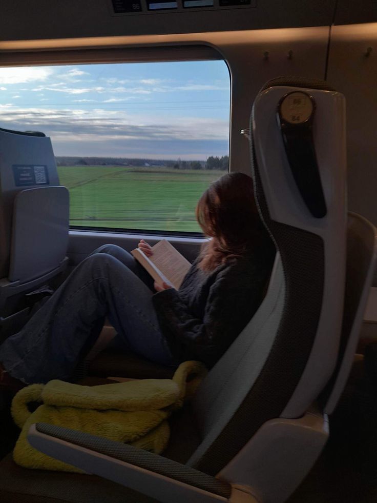 a woman sitting in an airplane looking out the window while reading a book on her lap