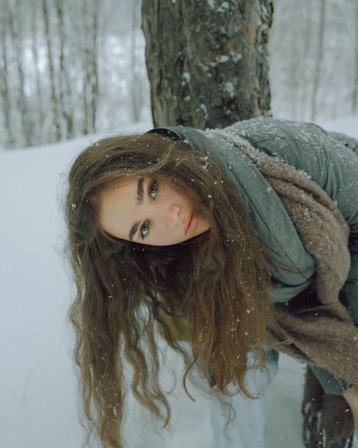 a woman with long hair standing next to a tree in the snow