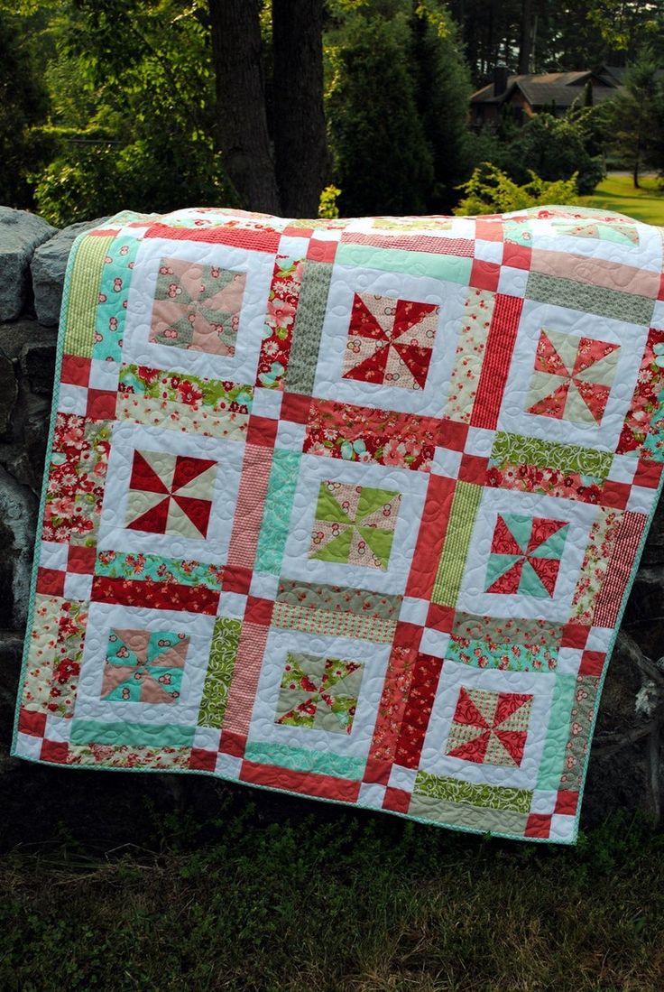 a red and green patchwork quilt hanging on a stone wall in front of some trees