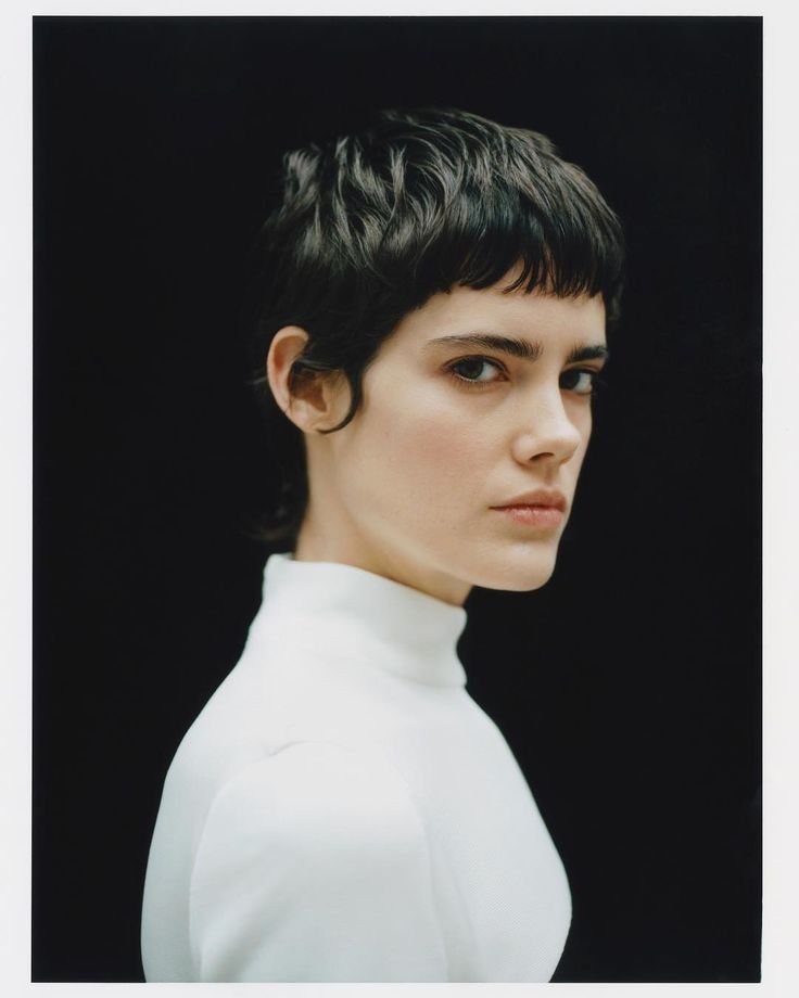 a woman with short black hair wearing a white shirt and looking at the camera while standing in front of a black background