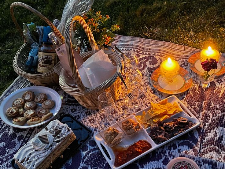 an outdoor picnic with food and candles on the blanket in the evening sun, next to two wicker baskets filled with snacks