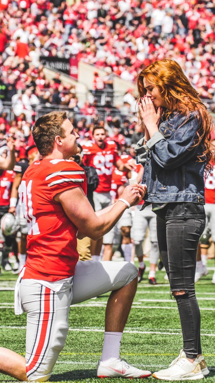 a man kneeling down next to a woman on top of a football field with fans in the background