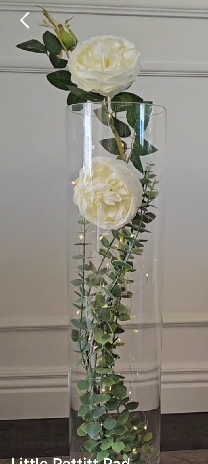 white roses in a clear vase with greenery and lights on the bottom, sitting on a wood floor