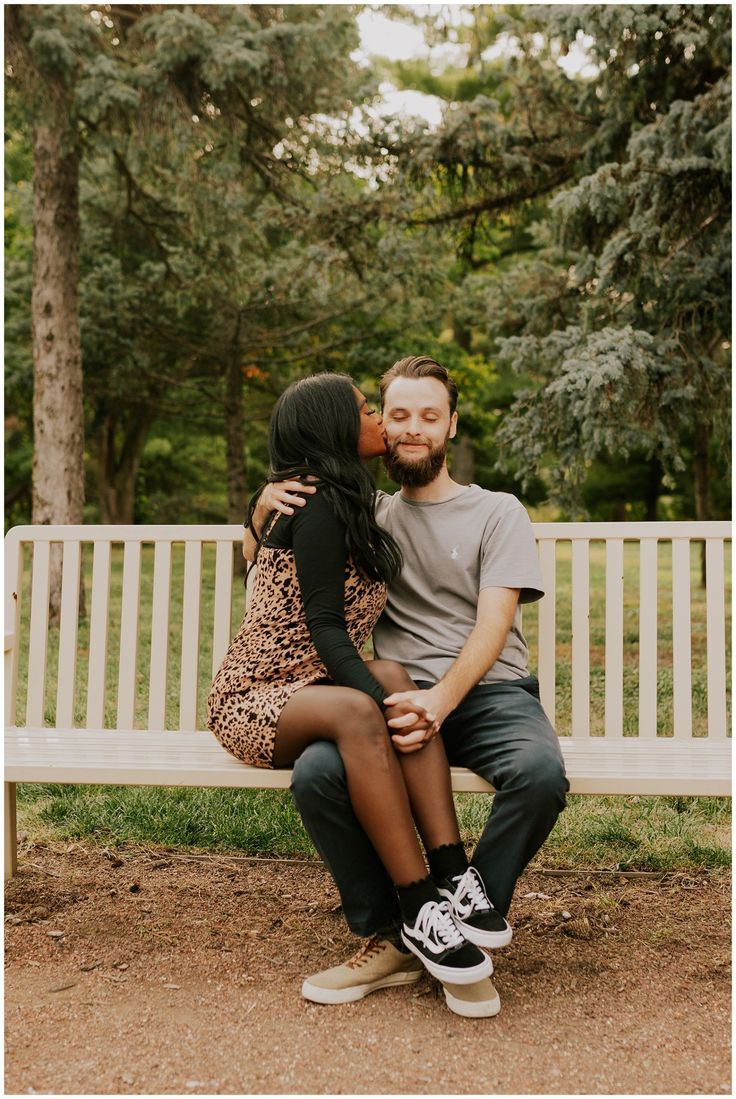 a man and woman sitting on a bench in front of some trees, one kissing the other