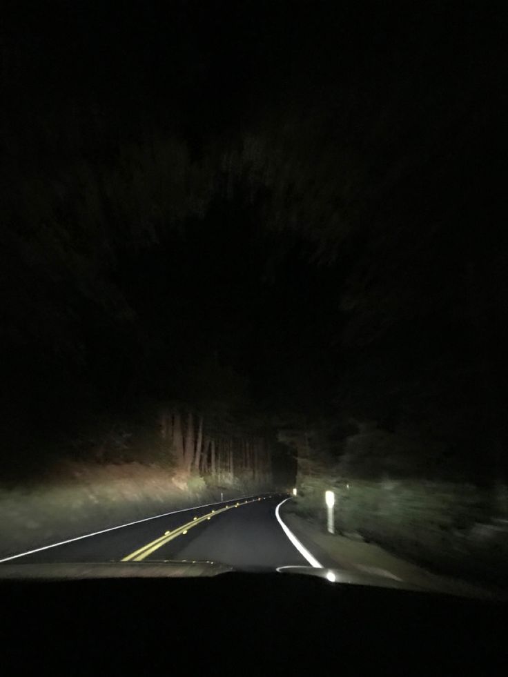 a car driving down a dark road at night with its headlights on and trees in the background