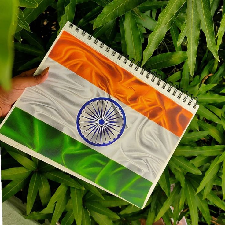 a hand holding a notebook with the flag of india on it in front of green leaves