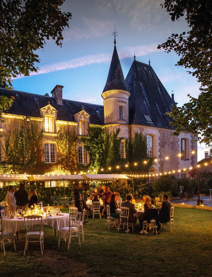 people sitting at tables in front of a large building with lights strung from the roof