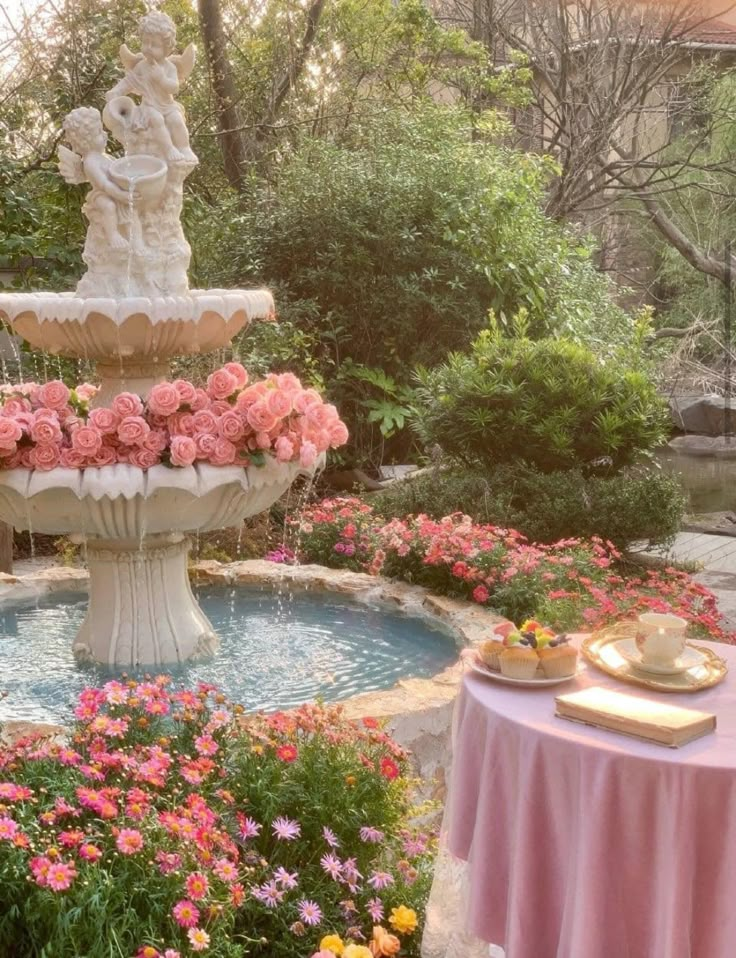 a fountain surrounded by pink flowers and other greenery in a garden area with tables