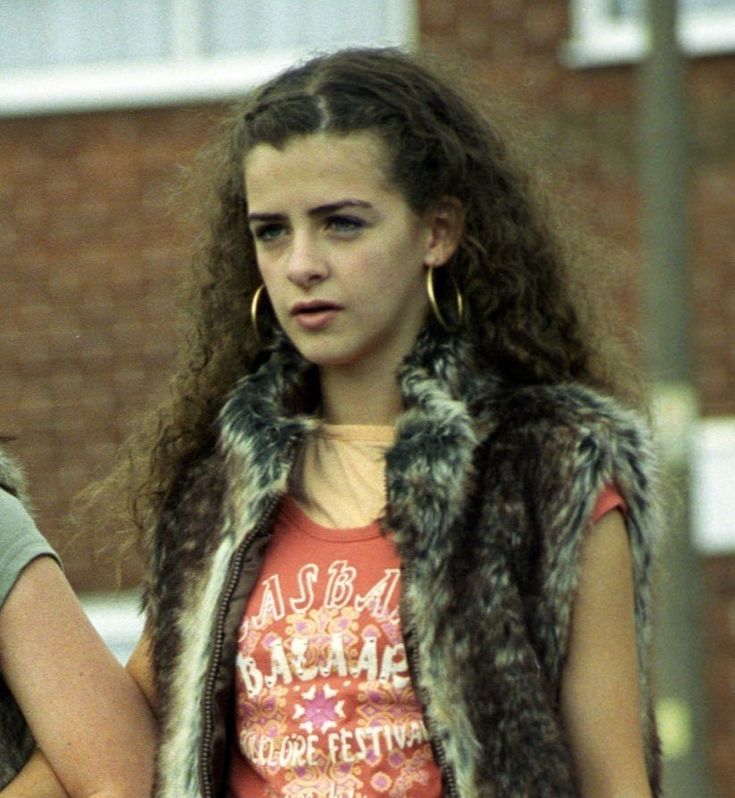 a girl with long hair wearing a fur vest and looking off to the side while standing in front of a brick building