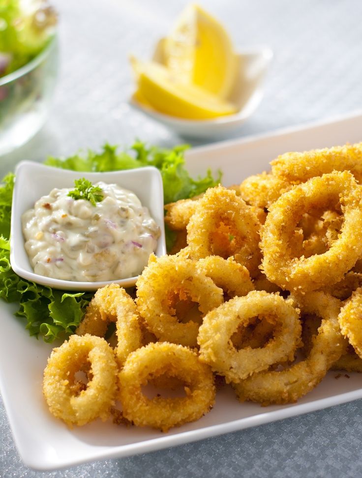 some onion rings are on a plate with dip and lemon wedges next to it