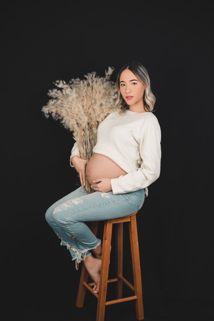 a pregnant woman sitting on a stool with dried grass in front of her belly and looking at the camera