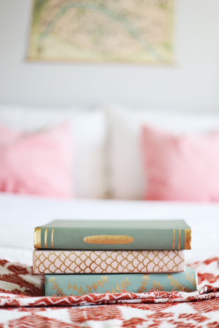 three books are stacked on top of each other in front of a bed with pink pillows