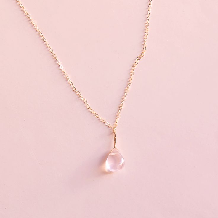 On a neutral background is a gold chain necklace with a rose quartz stone in the center. Rose Quartz Necklace In Rose Gold For Gift, Rose Gold Necklace With Rose Quartz As A Gift, Rose Gold Rose Quartz Necklace Gift, Feminine Rose Gold Rose Quartz Necklaces, Dainty Rose Gold Pendant Crystal Necklaces, Dainty Rose Quartz Pendant Jewelry, Minimalist Rose Quartz Jewelry As Gift, Minimalist Rose Quartz Jewelry For Gifts, Minimalist Rose Quartz Gemstone Jewelry