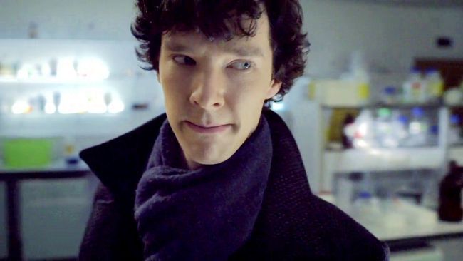 a man with curly hair wearing a black coat and scarf in a science lab looking at the camera