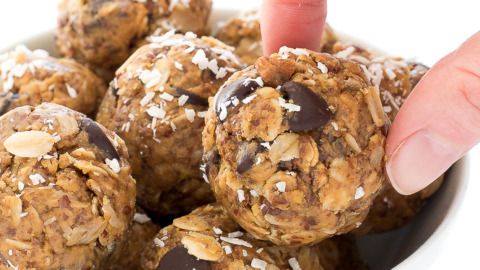 a hand picking up a chocolate chip cookie ball from a white bowl filled with oatmeal balls