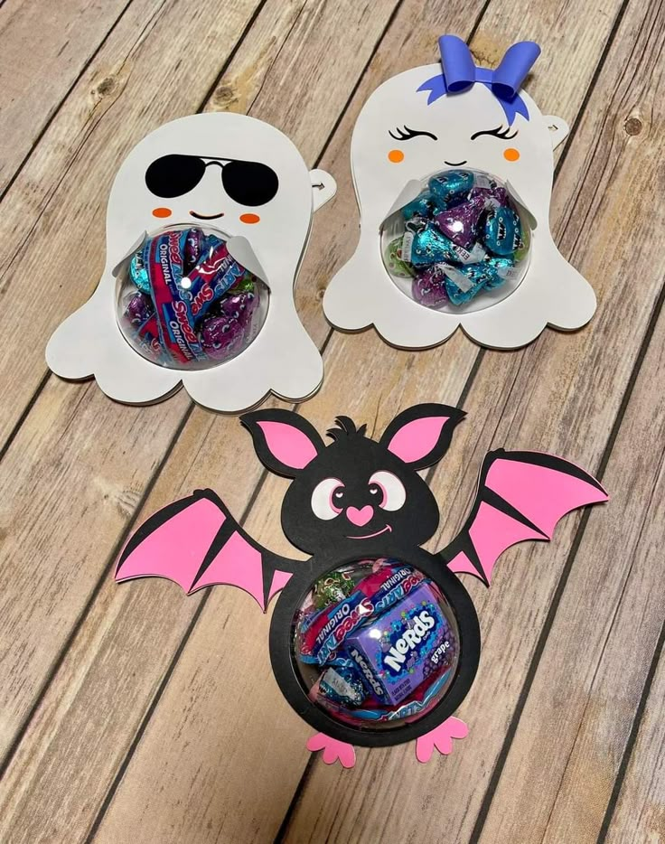 two halloween treat boxes with candy in them on a wooden table, one has a bat and the other has a ghost