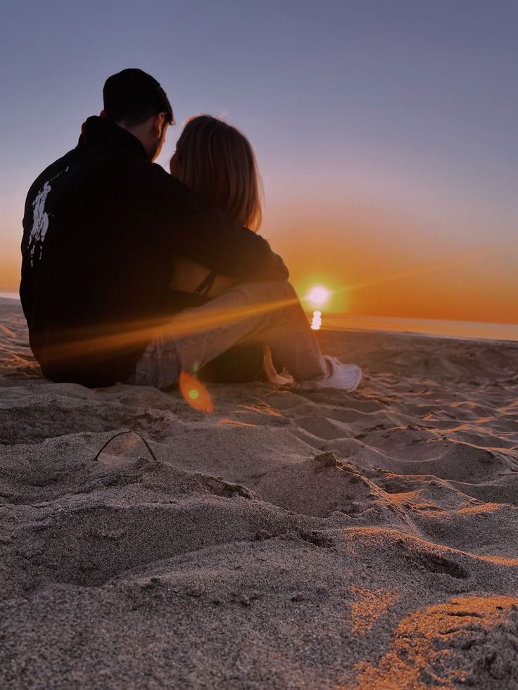 a man and woman sitting on the beach watching the sun go down over the ocean