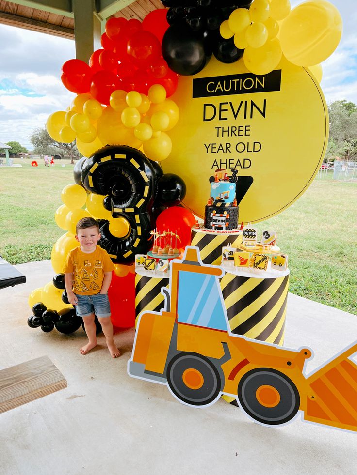 a young boy standing next to a construction themed birthday cake and balloon display at a children's birthday party