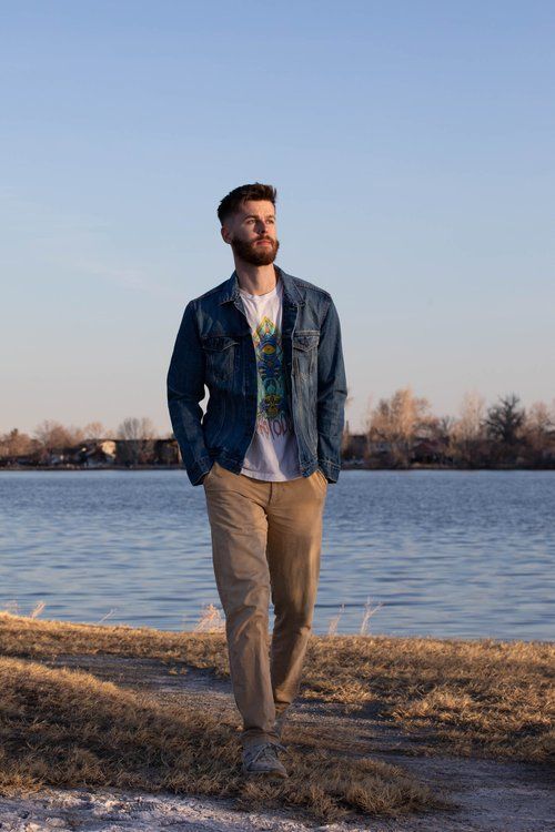 a man standing in front of a body of water wearing a denim jacket and khaki pants