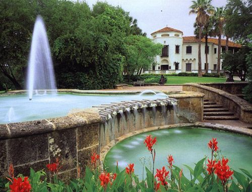 a fountain in the middle of a garden with red flowers around it and a large white building behind it