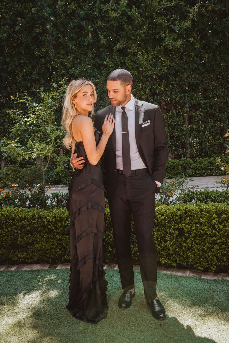 a man and woman standing next to each other in front of some bushes wearing formal clothes