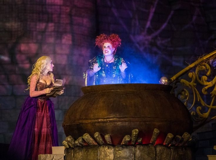 two women dressed in costumes standing next to a large pot