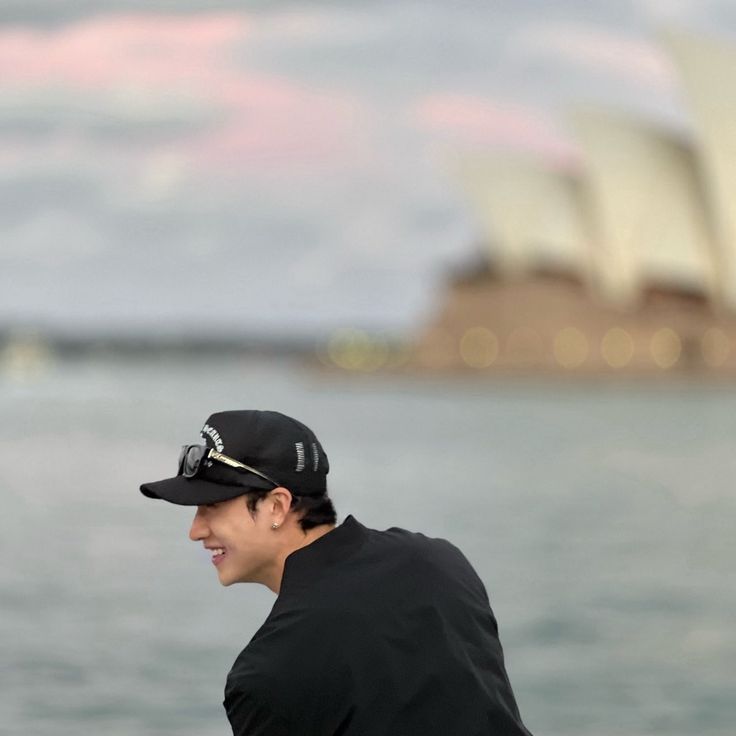 a man in black shirt and hat sitting next to the ocean with sydney opera house in background