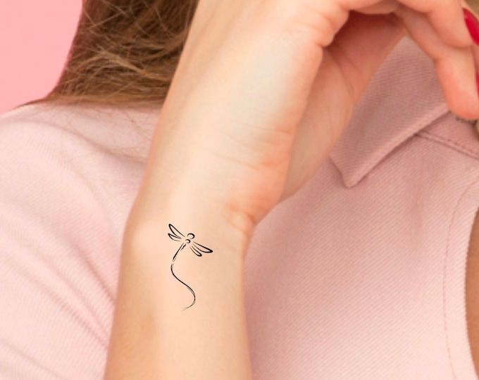 a woman with a small tattoo on her left wrist and hand behind her ear, she is wearing a pink shirt