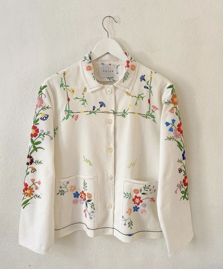 a white jacket with colorful flowers on it hanging from a hanger against a wall