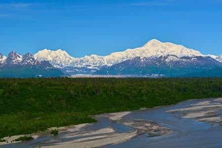 a river running through a lush green forest filled with snow covered mountains in the distance