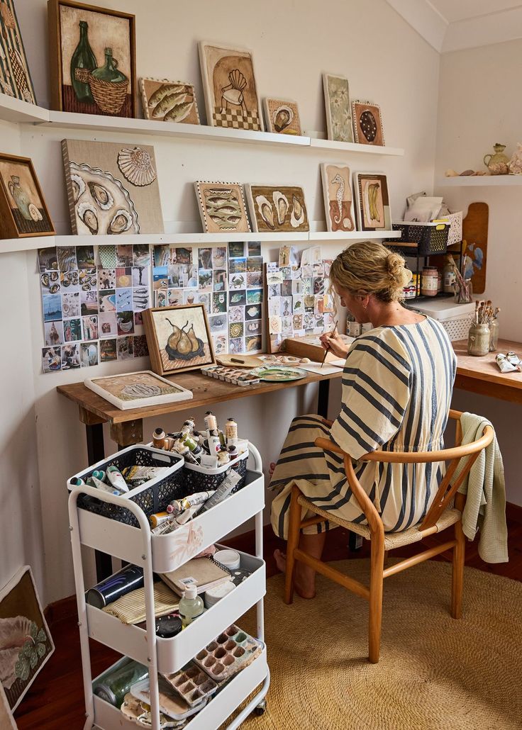 a woman sitting at a desk with lots of pictures on the wall behind her and she is painting