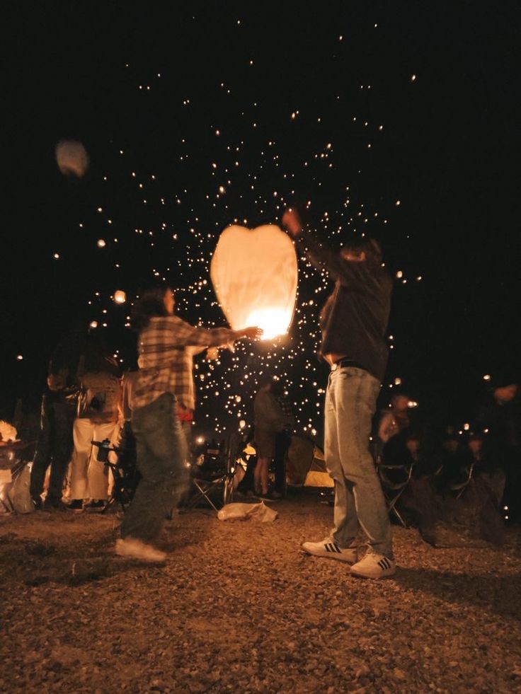 people standing around a fire in the dark with some lights on them and one person holding a paper lantern